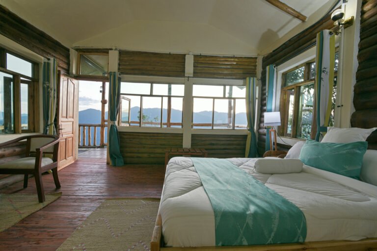 A lodge room with wooden walls, a large bed and windows onto forested hills