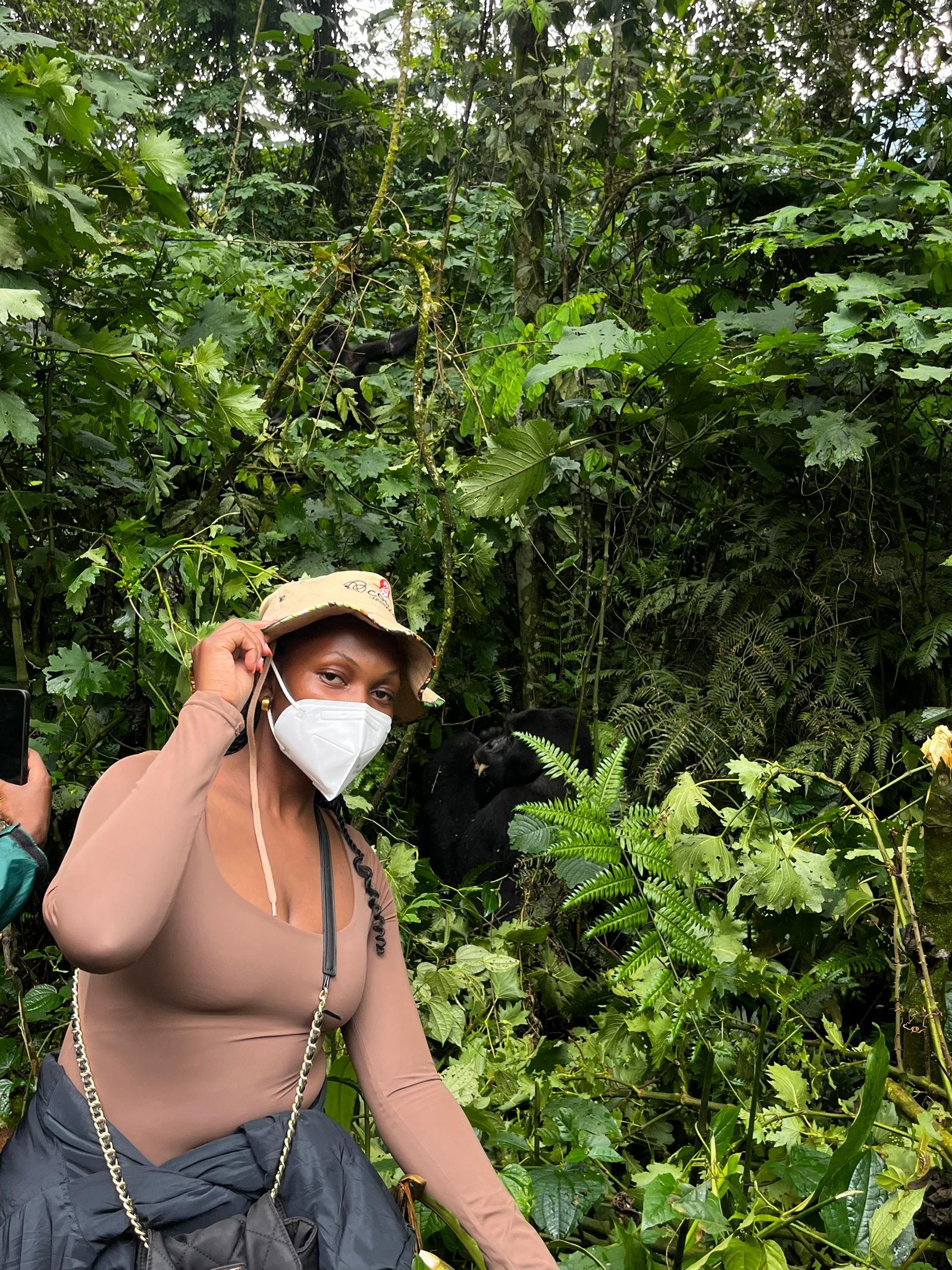 A guest on an Orugano Safaris gorilla trekking experience in Uganda’s Bwindi Impenetrable Forest, wearing a mask for safety, stands in lush rainforest foliage with a mountain gorilla visible close by, showcasing our close, responsible wildlife encounters led by local guides.